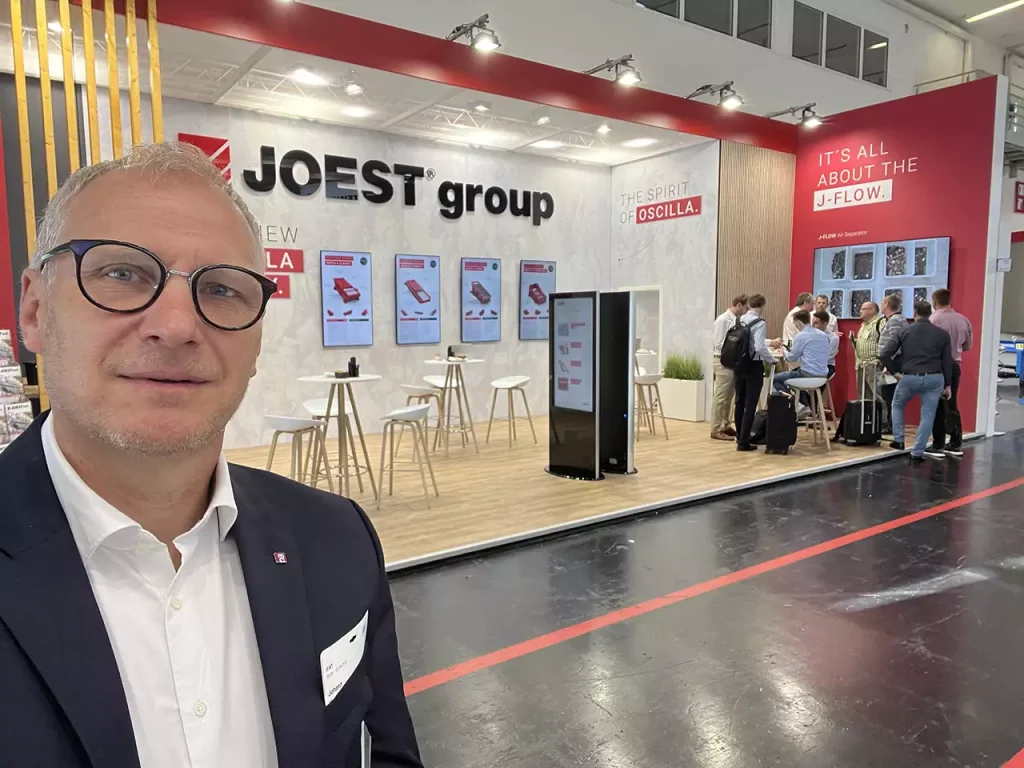 Mr. DI Johann Hagenauer with suit takes a selfie in front of the booth of JÖST. A few people can be seen at the bar tables in front of the presented pictures